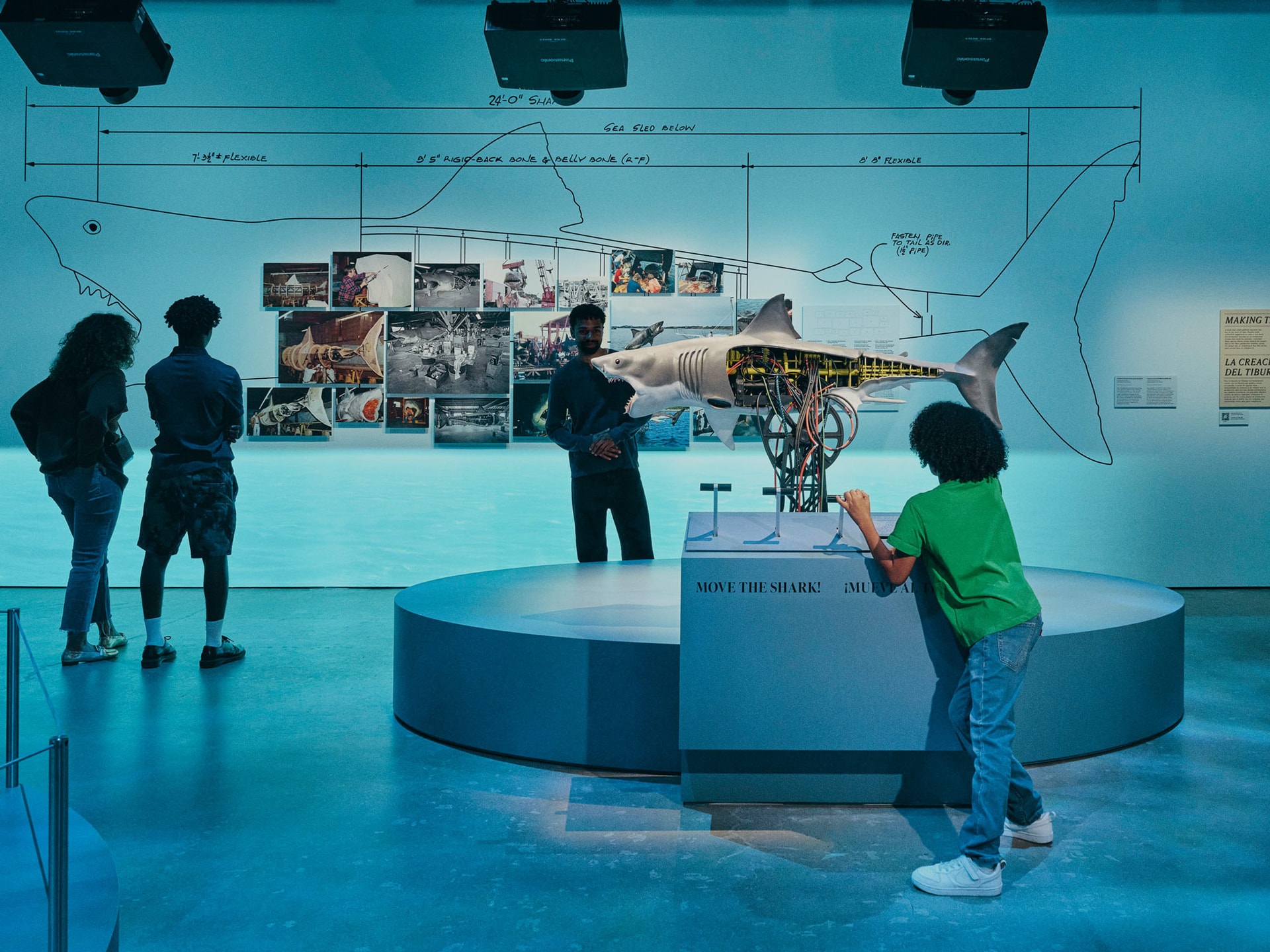 An image from an exhibit at the Academy Museum of Motion Pictures. The display is set in a dark room illuminated by blue lighting. The main wall features a large, schematic line drawing of a great white shark. Overlaid on the drawing is a collage of behind-the-scenes photographs related to the film Jaws and a three-dimensional, partially sectioned model of the mechanical shark, "Bruce," revealing its internal gears and mechanics. Three adult visitors stand on the left, looking at the display. In the foreground, a young boy in a green shirt and jeans is interacting with a control panel on a circular, multi-tiered pedestal in front of the model. The pedestal has text that reads, "MOVE THE SHARK! ¡MUEV..."