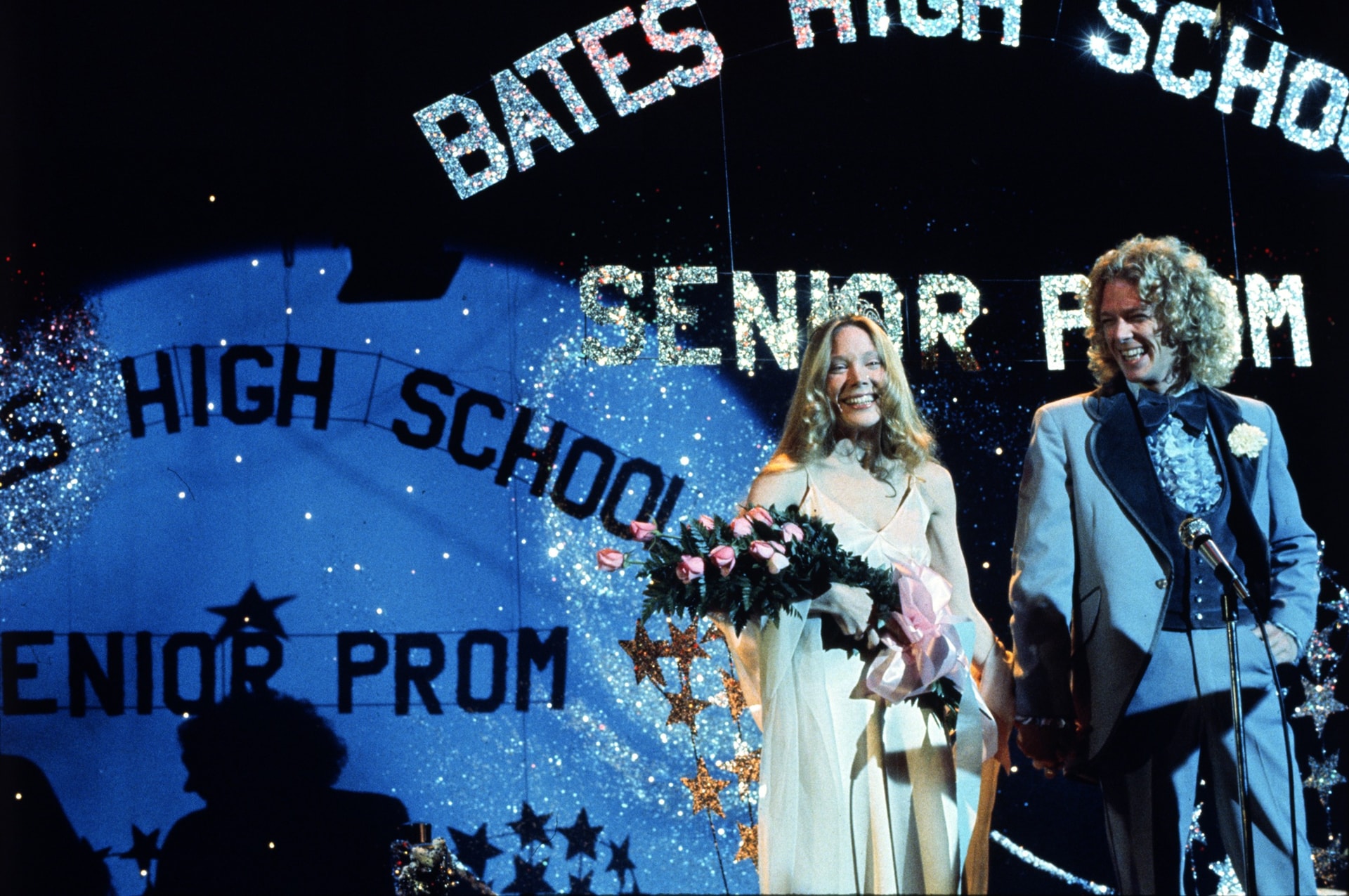 Sissy Spacek as Carrie White, crowned prom queen in a white dress holding a bouquet of pink roses, stands beside William Katt as Tommy Ross under a glittering 'Bates High School Senior Prom' sign in a still from Carrie (1976).
