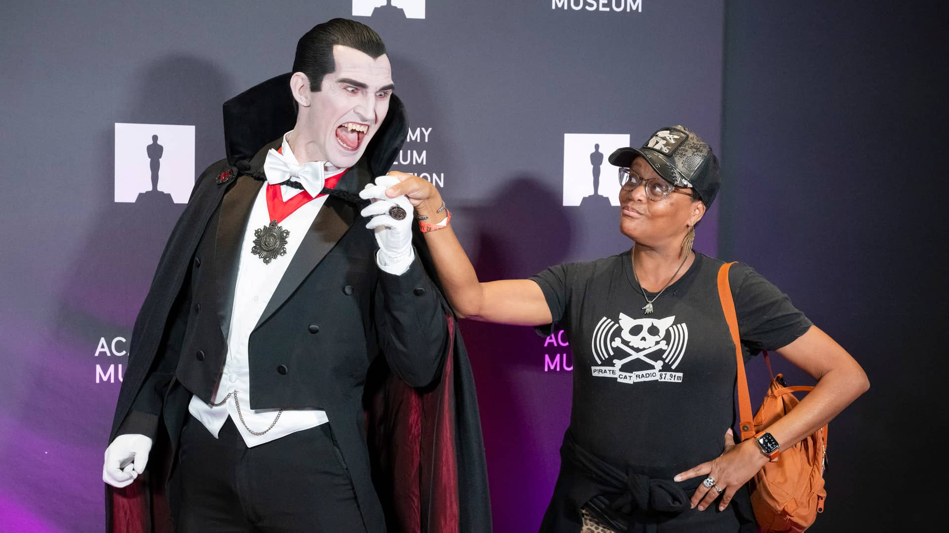 At the Academy Museum's Monster Mash event, a woman in a Pirate Cat Radio t-shirt playfully touches the cape of a costumed Dracula performer.